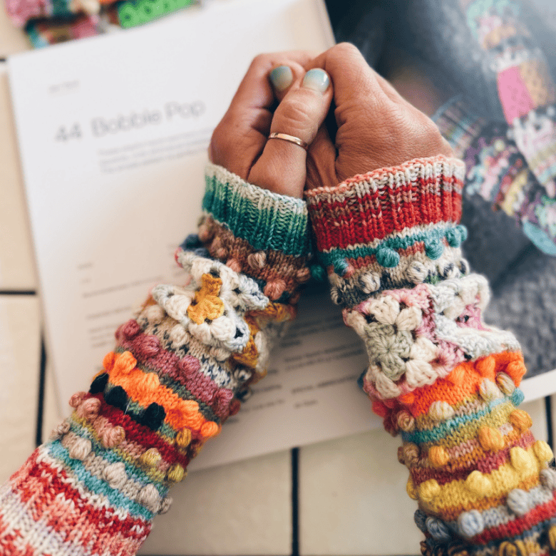 A close-up shows a person with medium skin tone clasping their hands together over an open book, wearing colorful, hand-knit fingerless gloves decorated with stripes, bobbles, and small crocheted flowers; the person has short, painted nails in a muted green shade and a simple ring on one finger, while the softly blurred background suggests a cozy indoor setting focused on knitting or crafting.