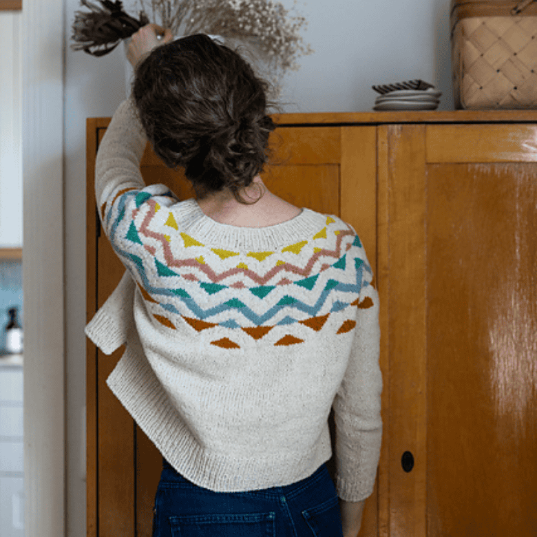 A person with light skin tone is seen from behind indoors, wearing a cream-colored knitted sweater with colorful geometric patterns across the shoulders, reaching up to arrange dried flowers on top of a wooden cabinet in a cozy home setting.