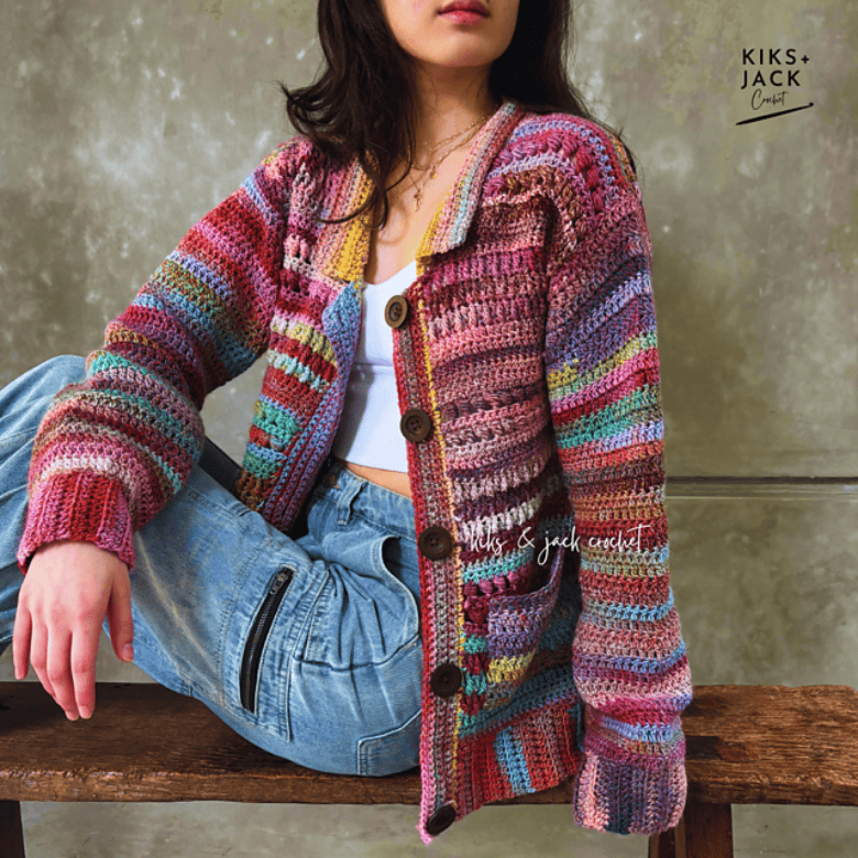 A seated person with medium-light skin tone wears a multicolored striped crocheted cardigan over a white top and blue jeans, posing casually on a wooden bench against a neutral backdrop, with the garment’s texture and large buttons clearly visible.