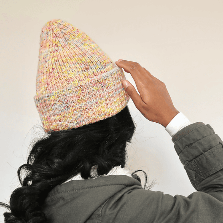 A person with medium-dark skin tone is shown from behind adjusting a speckled, pastel-colored knitted beanie on their head, with long dark hair visible beneath the hat and a neutral indoor background.