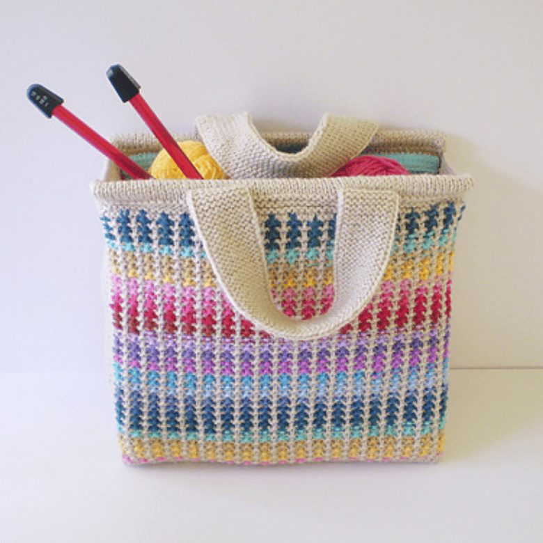A rectangular knitted tote bag with multicolored stripes and cream handles sits against a plain background, filled with yarn and knitting needles, showcasing a handmade craft item; no people are visible.