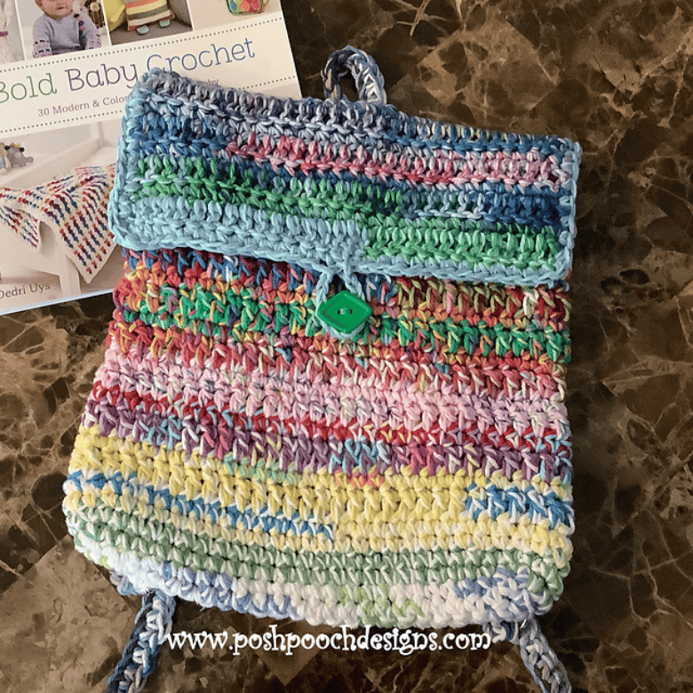 A colorful crocheted pouch or small bag with horizontal stripes lies on a marble surface next to a book titled “Bold Baby Crochet,” highlighting a handmade accessory; no people are visible.