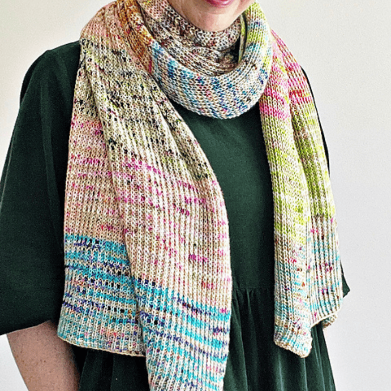 A person with light skin tone models a long, multicolored knitted scarf draped around their neck over a dark green top, with only the lower half of their face visible and a neutral background.