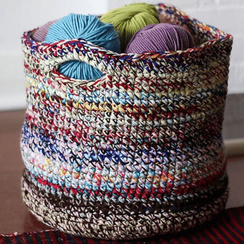 A round, multicolored crocheted basket filled with balls of yarn sits on a wooden surface, emphasizing texture and color variation; no people are visible.