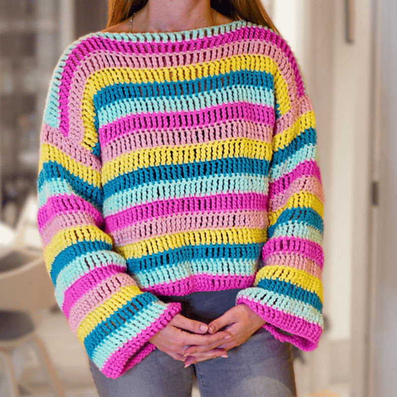 A person with light skin tone wears a brightly striped crocheted sweater in pink, yellow, teal, and purple hues, standing indoors with hands clasped in front, focusing on the garment’s bold pattern.