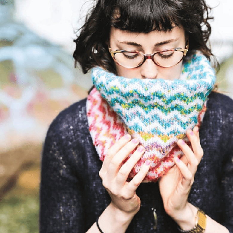 A person with light skin tone wearing glasses holds a thick, multicolored knitted cowl or scarf up to their face outdoors, with short dark hair and a softly blurred background.