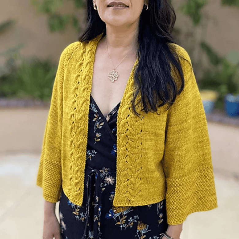 A woman with a medium-light skin tone is shown from the neck down, wearing a bright mustard-yellow knitted cardigan featuring an open front with decorative cable stitch borders and wide, loose-fitting sleeves. She wears the cardigan over a black floral-patterned dress, accessorized with a delicate flower pendant necklace. Her long, dark hair falls over her left shoulder against a softly blurred outdoor garden backdrop.