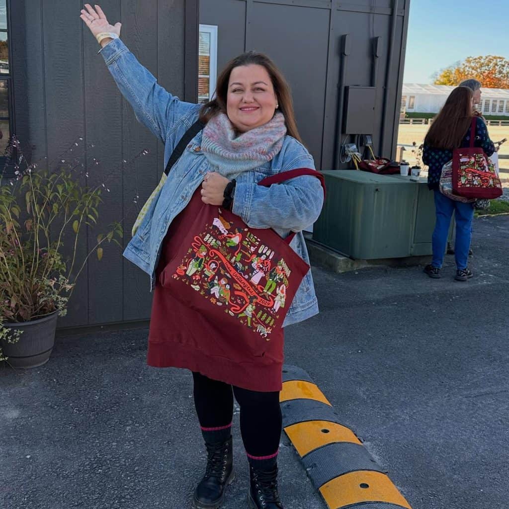 A smiling woman with a medium-light skin tone stands outdoors on asphalt, waving her right arm. She wears a denim jacket, maroon dress, multicolored knit scarf, black leggings, and combat boots while holding a maroon tote bag that reads "Indie Untangled 2024." In the background, near a dark grey building, another woman with a light skin tone is seen from behind wearing blue jeans and a patterned red tote bag.