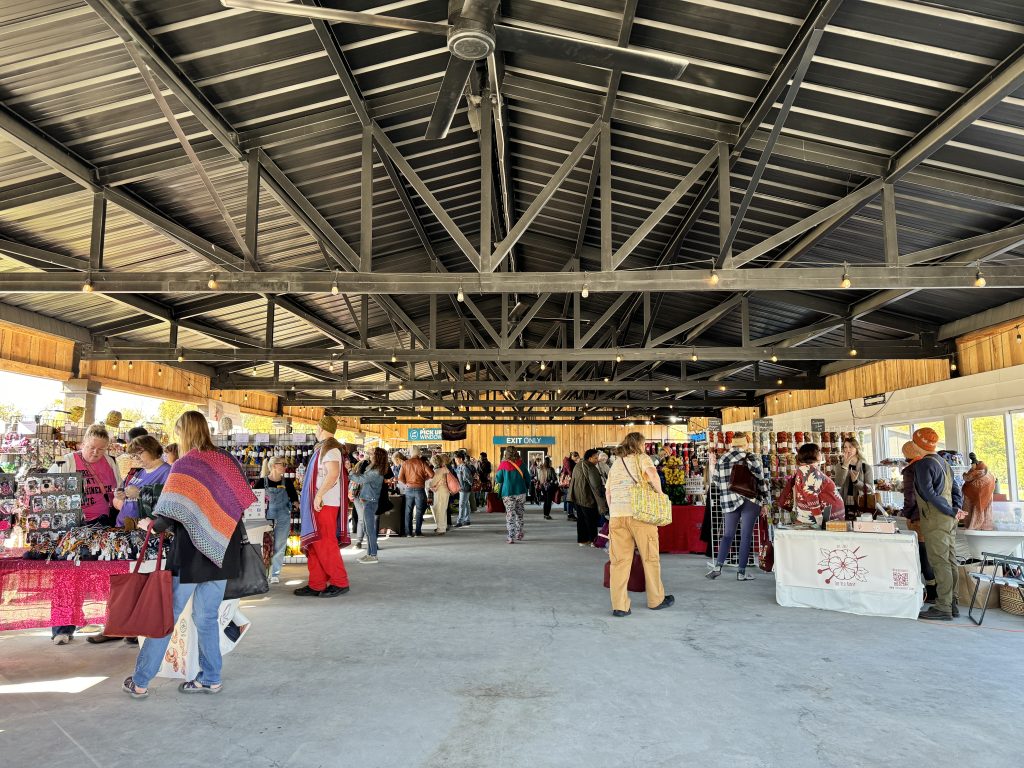 A spacious, open-air pavilion with a high, vaulted metal roof shelters a bustling craft market. Scattered across the concrete floor, shoppers browse booths filled with colorful yarn, clothing, and handmade crafts beneath strings of warm bulb lights. In the left foreground, several shoppers with light skin tones look at a display table, including a woman in a vibrant, multicolored knit shawl, while other market-goers with light skin tones, medium-light skin tones, and medium-dark skin tones mill about the wide central aisle.