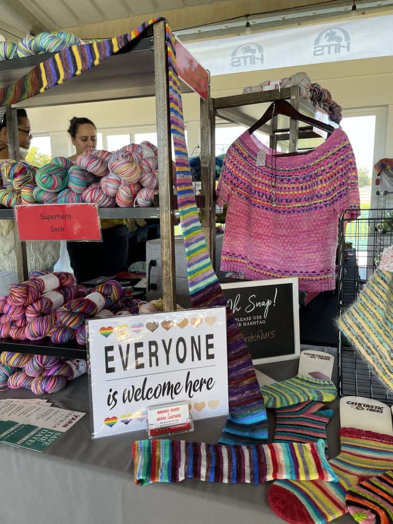 A vendor booth displays vibrant hand-dyed yarn and knitwear under an outdoor pavilion. In the foreground, a sign reads "EVERYONE is welcome here" alongside multicolored striped socks, a long striped scarf, and shelves filled with bright yarn skeins labeled "Superhero Sock." To the right hangs a pink knitted sweater with a patterned yoke, while a woman with a medium skin tone and a woman with a light skin tone are visible in the blurred background looking at the merchandise.