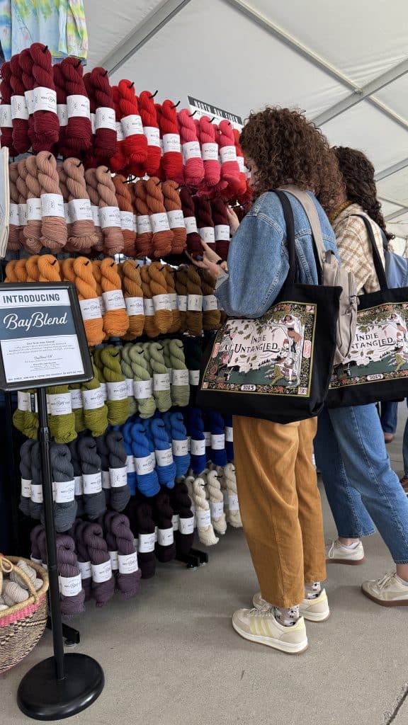 Two women with light skin tones stand in profile inside a tented market booth, browsing a large vertical wall display of colorful yarn skeins arranged in a gradient. The woman in the foreground has long, curly brown hair, wears a denim jacket and mustard corduroy pants, and reaches out to touch a dark red skein. Both women carry matching black canvas tote bags that read "Indie Untangled 2025," and a sign in the lower left introduces "BayBlend" yarn.