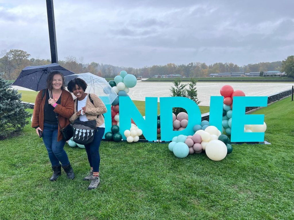 Two smiling women stand outdoors under umbrellas on a green lawn next to a large teal sign reading "INDIE," which is decorated with colorful balloons. The woman on the left has a light skin tone and wears a rust-colored cardigan, while the woman on the right has a medium-dark skin tone and wears a beige knit sweater. An open sandy track and a distant treeline sit beneath an overcast sky in the background.