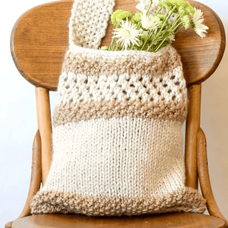A textured, hand-knitted tote bag hangs over the back of a vintage wooden chair. The bag features alternating horizontal bands of cream and light brown yarn, incorporating a decorative openwork diamond stitch pattern near the top. Green and white flowers peek out from the open top of the tote against a plain white background.