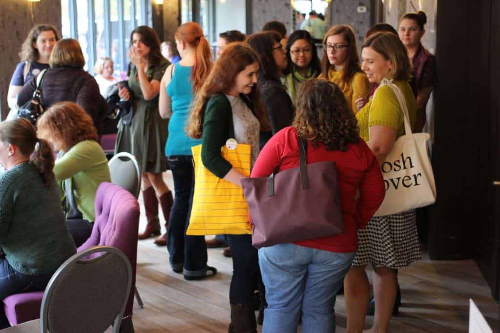 A group of people are gathered in a well-lit, narrow indoor venue with light wood floors. In the foreground, several individuals with light skin tone are chatting, including a woman in a red top with a purple tote bag seen from behind, a smiling woman facing her with long brown hair and a yellow tote, and a woman in a yellow top holding a canvas bag. To the left, others with light skin tone sit on purple upholstered chairs or stand near the windows. In the background, additional attendees with light skin tone and medium-light skin tone stand in small clusters, creating a lively social gathering.