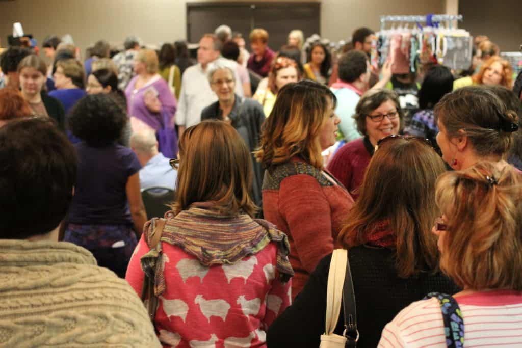 A crowded indoor gathering features a tight assembly of people, primarily with light skin tone and medium-light skin tone, milling around a market or convention space. In the foreground, several individuals are seen from behind, including one in a textured beige knit sweater and another in a pink sweater patterned with white sheep. The bustling background is filled with dozens of other attendees chatting and browsing displays under warm indoor lighting.