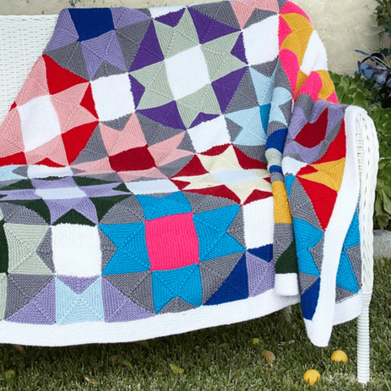 A vibrant, multicolored knit blanket is draped over a white wicker outdoor sofa sitting on green grass. The blanket features a complex geometric patchwork pattern of colorful knit stars and squares in shades of red, blue, purple, magenta, yellow, teal, and grey, all framed by a clean white border.