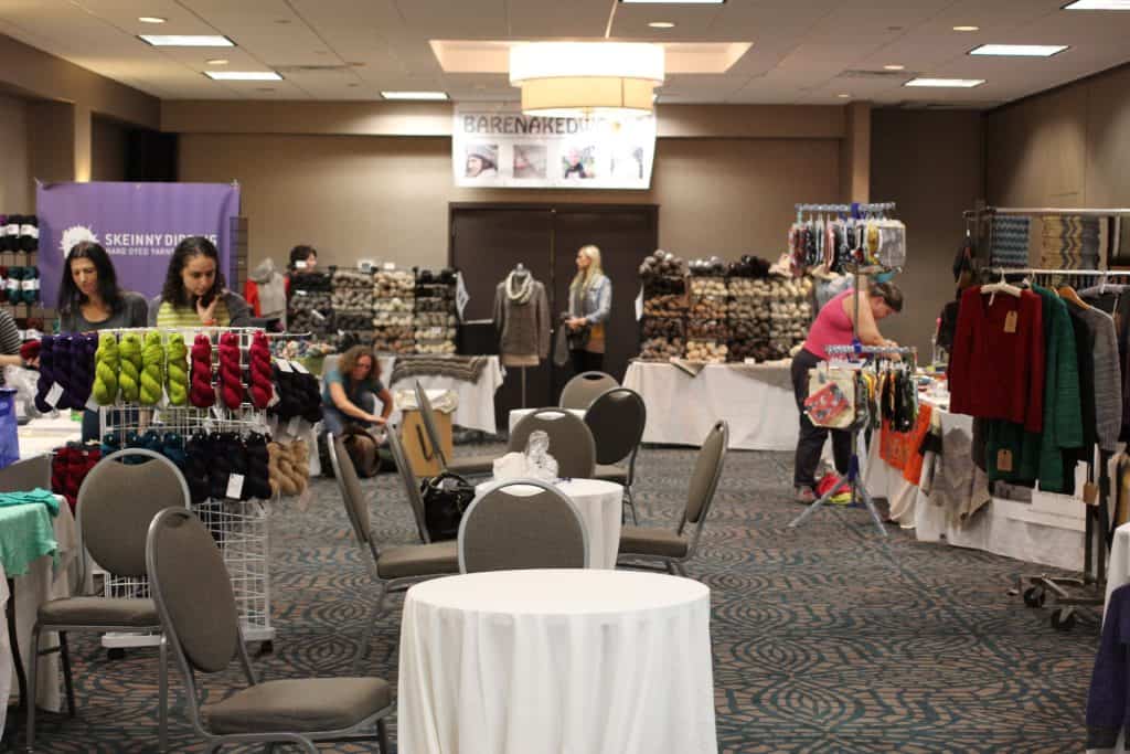 A large indoor yarn and knitting market is set up in a hotel conference room with tables, chairs, and vendor booths displaying colorful skeins of yarn and handmade clothing. Several people with light skin tone and medium-light skin tone browse the displays, talk with vendors, and arrange merchandise under warm overhead lighting.