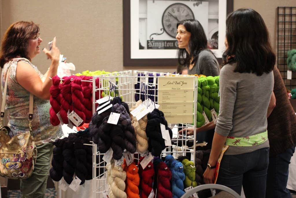 Three women stand around a display rack filled with brightly colored yarn bundles at a craft fair booth. The women appear to have light skin tone and medium-light skin tone, and they are talking while looking at the yarn display in a cozy indoor event space.