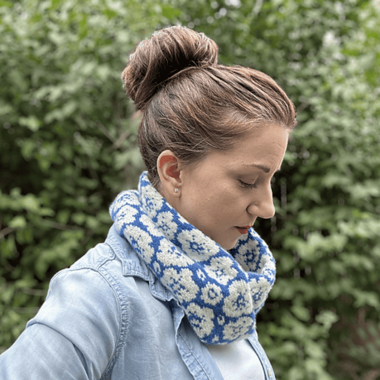 A woman with a light skin tone is shown in profile, looking down while wearing a thick, cowl-style knit scarf with a blue and white floral pattern. Her hair is styled in a high bun, and she wears a light-wash denim button-down shirt over a white top against a blurred background of green foliage.