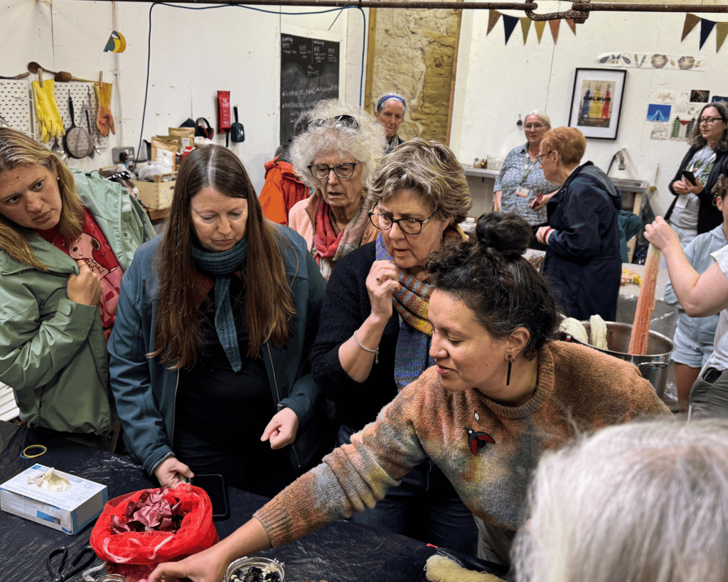 A group of women with light and medium skin tones crowd around a workbench in a rustic studio, intently watching a natural dyeing demonstration with plant materials and bowls.