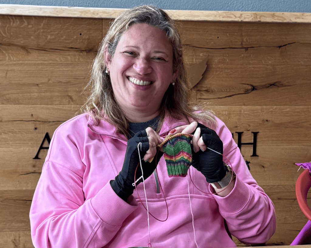 A woman with medium-light skin tone in a pink hoodie laughs brightly while knitting a small rainbow-striped project, seated in front of a wooden plank wall.