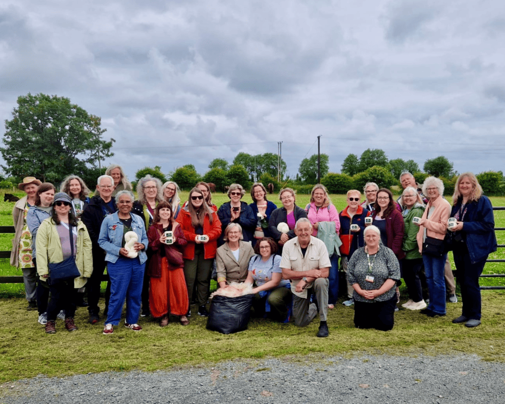 A large group of about 25 people with a mix of light, medium-light, medium, and dark skin tones smile for a photo in a green farm field under a cloudy sky, many holding small white wool bundles.