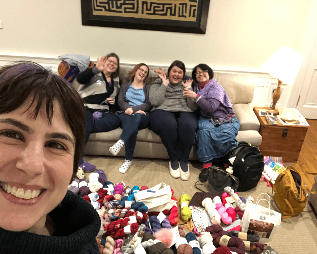 A selfie of five women with light and medium-light skin tones laughing in a living room surrounded by a large pile of colorful yarn skeins on the floor.