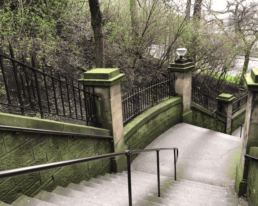Stone steps with moss-covered sandstone walls and black iron railings descending through a wooded area with bare early-spring trees.
