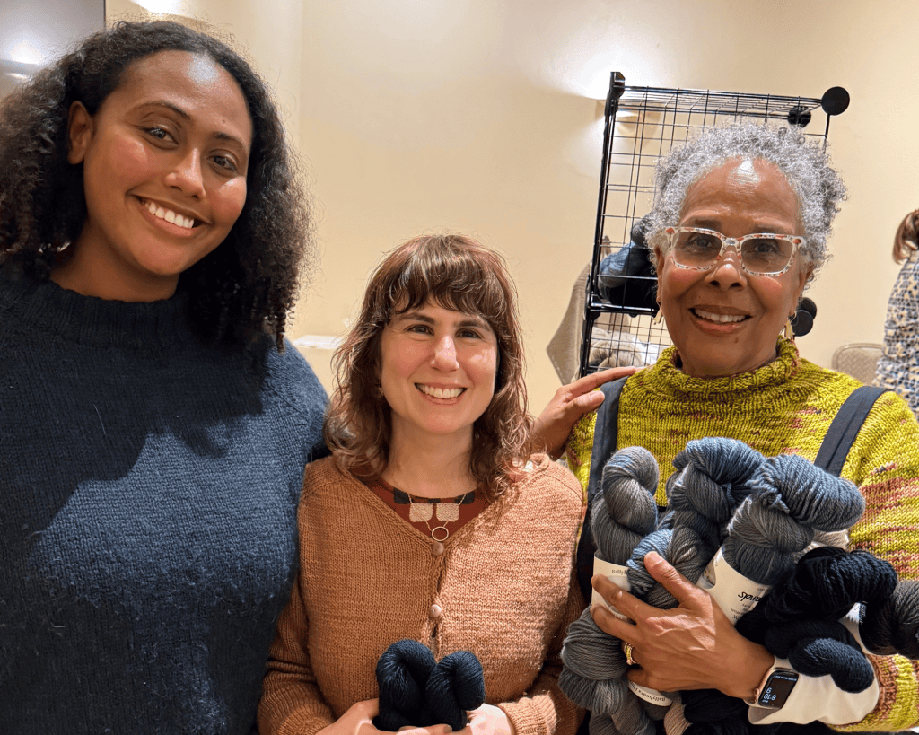 Three women with medium-dark, medium-light, and dark skin tones smile together, two holding skeins of blue-gray yarn, in a warmly lit room.
