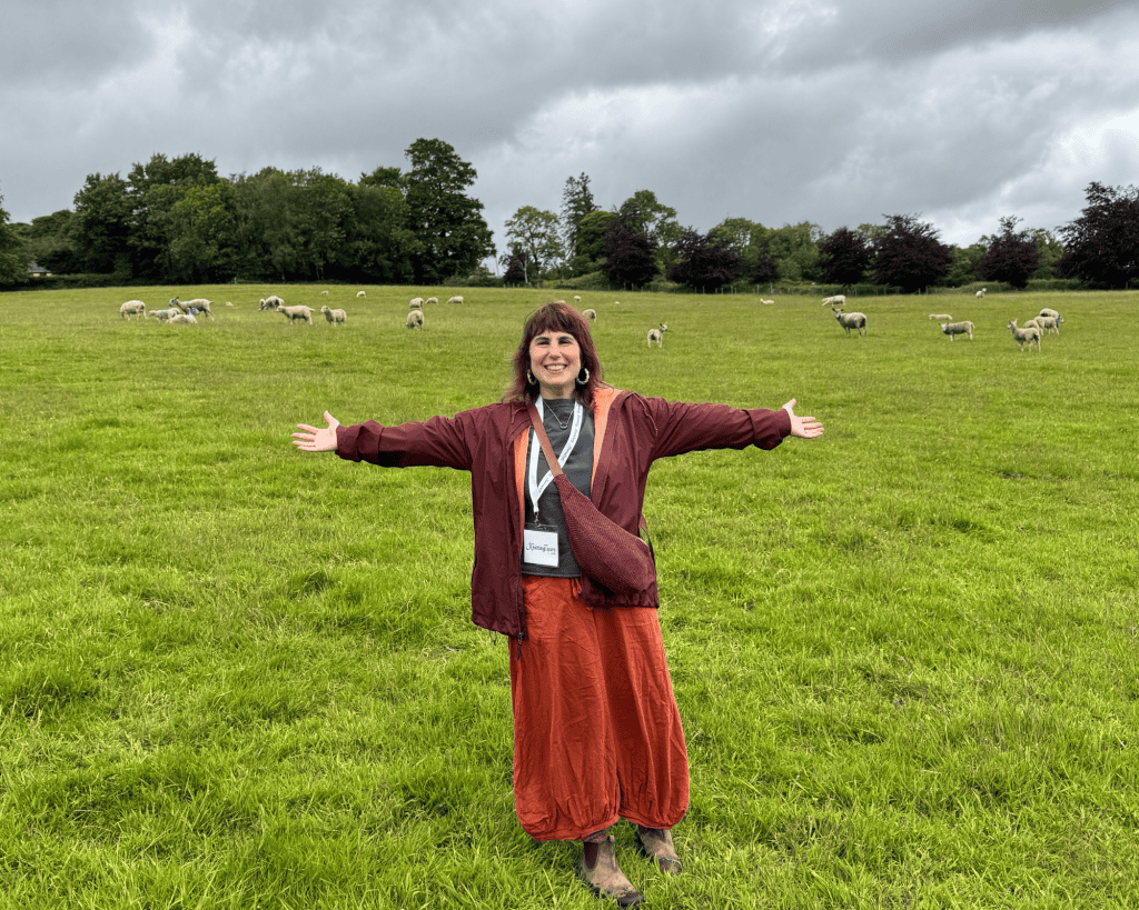 A woman with medium-light skin tone in a burgundy jacket and orange skirt stands arms outstretched, smiling in a green sheep pasture under a cloudy sky.