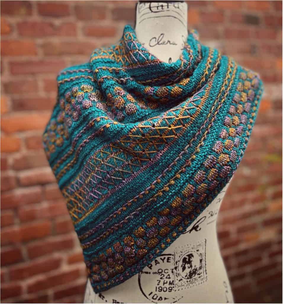 A teal, gold, and purple patterned knit shawl draped on a fabric mannequin in front of a brick wall.