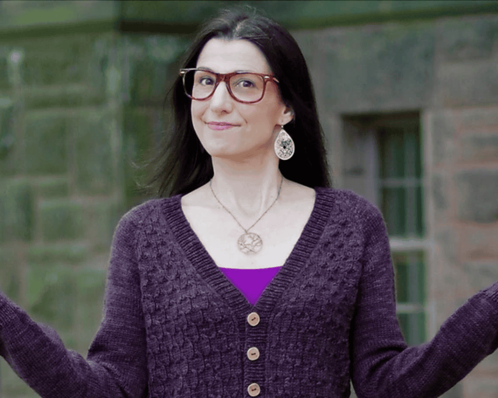 A woman with light skin tone and long dark hair, wearing large red-framed glasses and dangling earrings, smiles slightly with arms outstretched, dressed in a purple cable-knit cardigan over a magenta top, standing in front of a stone building.