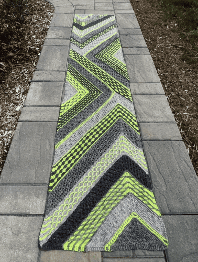 A long, colorblocked knit scarf in gray, black, and neon green with zigzag stitch patterns, laid out on a stone pathway.