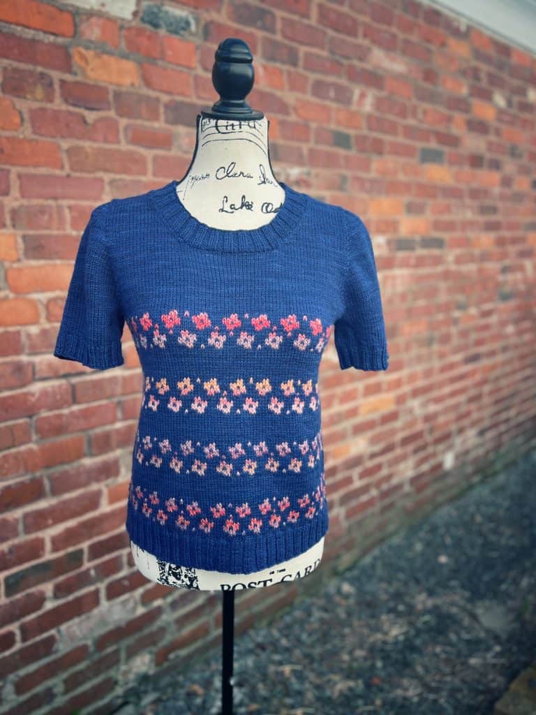 A navy blue short-sleeve knit sweater with rows of small pink and orange floral motifs, displayed on a fabric mannequin against a brick wall.