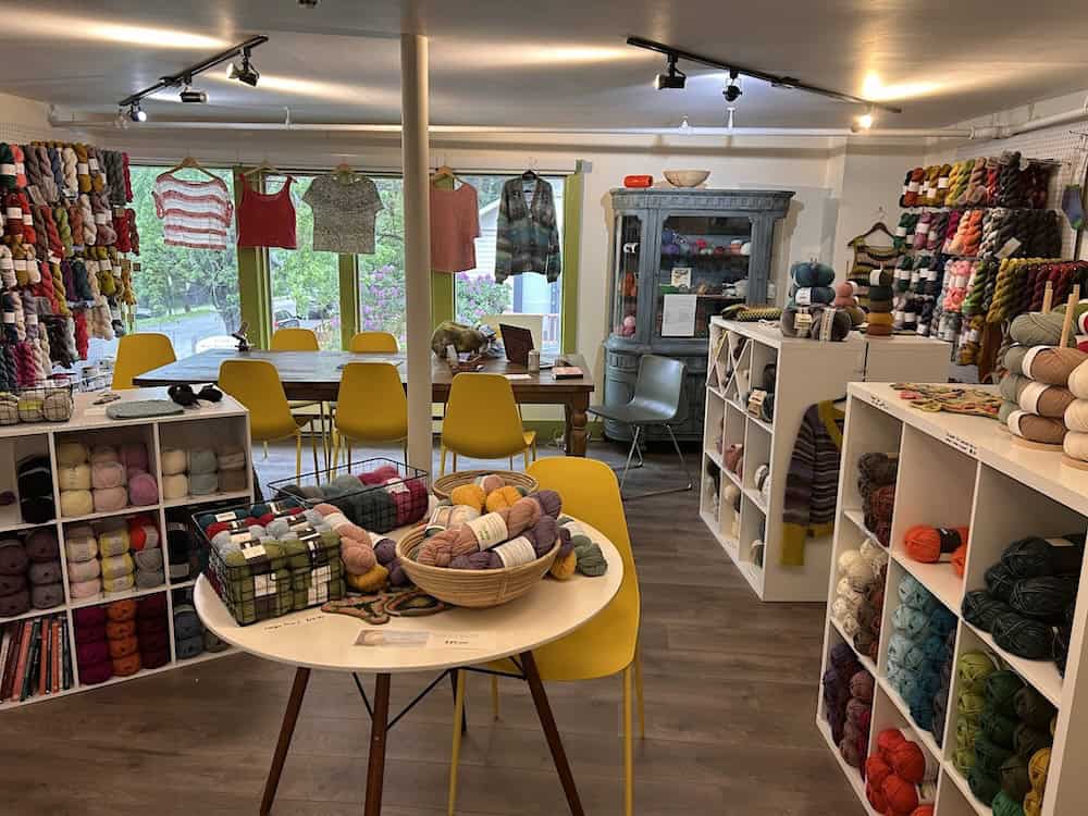 Interior of a yarn shop with walls of colorful yarn skeins, white cubby shelving, a wooden table with yellow chairs, knit garments hanging by the window, and a display table with baskets of yarn.