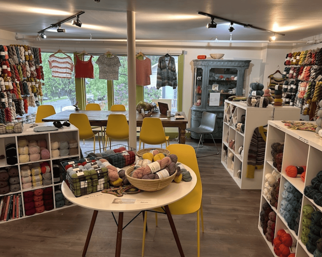 Interior of a yarn shop with walls of colorful yarn skeins, white cubby shelving, a wooden table with yellow chairs, knit garments hanging by the window, and a display table with baskets of yarn.