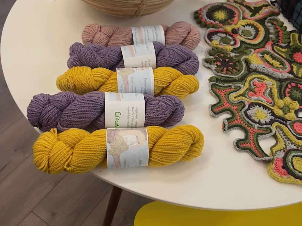A round white table displaying skeins of pink, purple, and mustard yellow yarn beside a colorful crocheted flower motif piece.