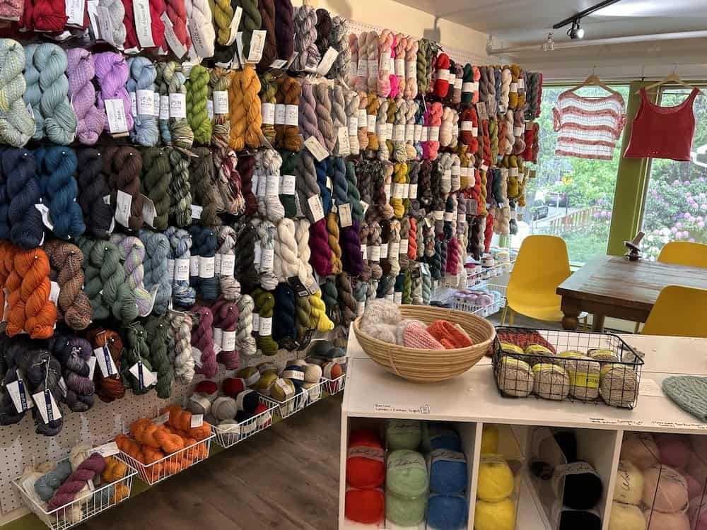 A yarn shop wall densely covered with rows of colorful hanging yarn skeins in blues, greens, pinks, and neutrals, with a display table, baskets of yarn, and knit sweaters visible near a window.