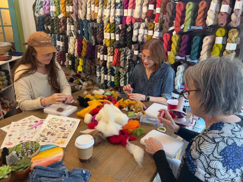 Three women with light skin tone sit around a wooden table felting wool in a yarn shop, surrounded by walls of colorful hanging yarn skeins.