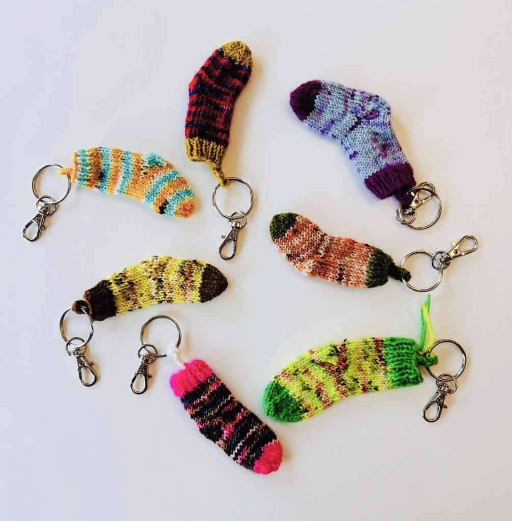 Six small knitted sock-shaped keychains in varied bright colors and stripe patterns, each attached to a keyring clip, arranged on a white surface.