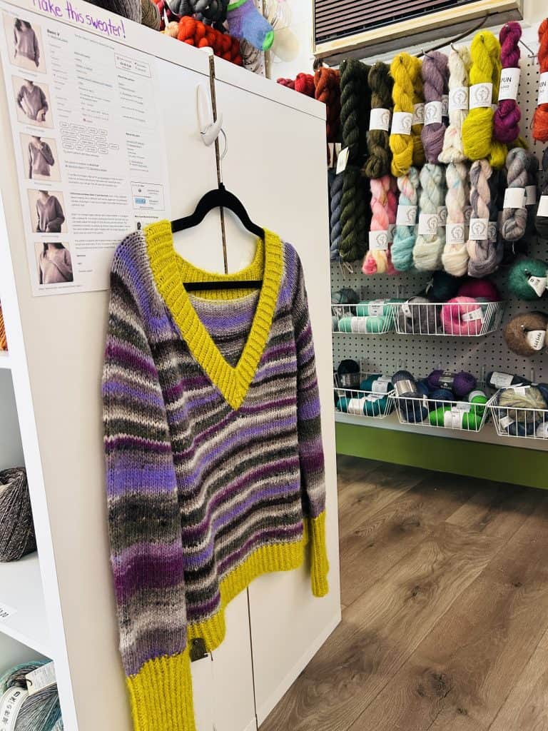 A purple, gray, and mustard yellow striped V-neck sweater hangs on a cabinet beside a printed pattern sheet, with a wall of colorful yarn skeins and baskets in the background.