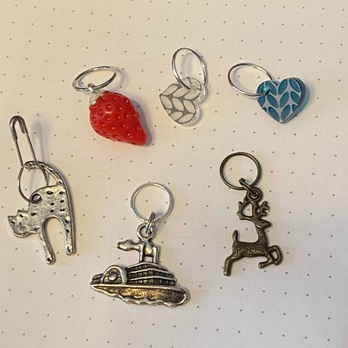 Set of 6 stitch markers: a silver cat, boat and stag, a strawberry and clear and blue hearts with stockinette stitch.