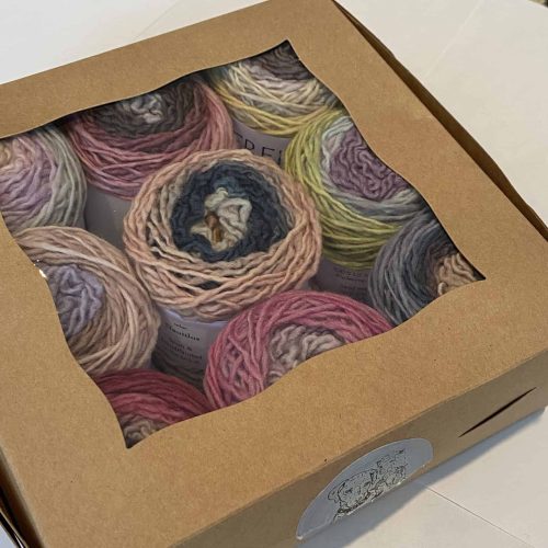 A set of 9 cakes of hand painted yarns in various muted ombre colors in a brown box with a clear window.