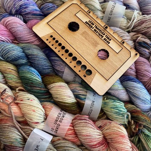 A wooden needle gauge in the shape of a cassette tape atop mini skeins of yarn in various bright colors.