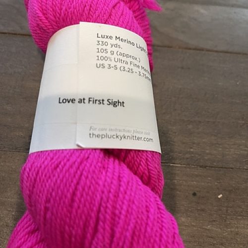 A skein of hot pink Plucky Knitter yarn with a label that reads Love at First Sight.